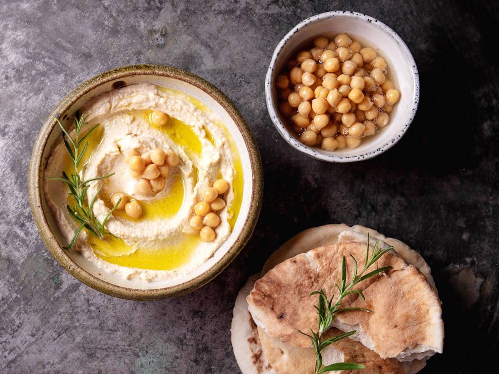 Kikerter i en liten skål, en skive brød med urter og en skål med hummus på en mørk overflate