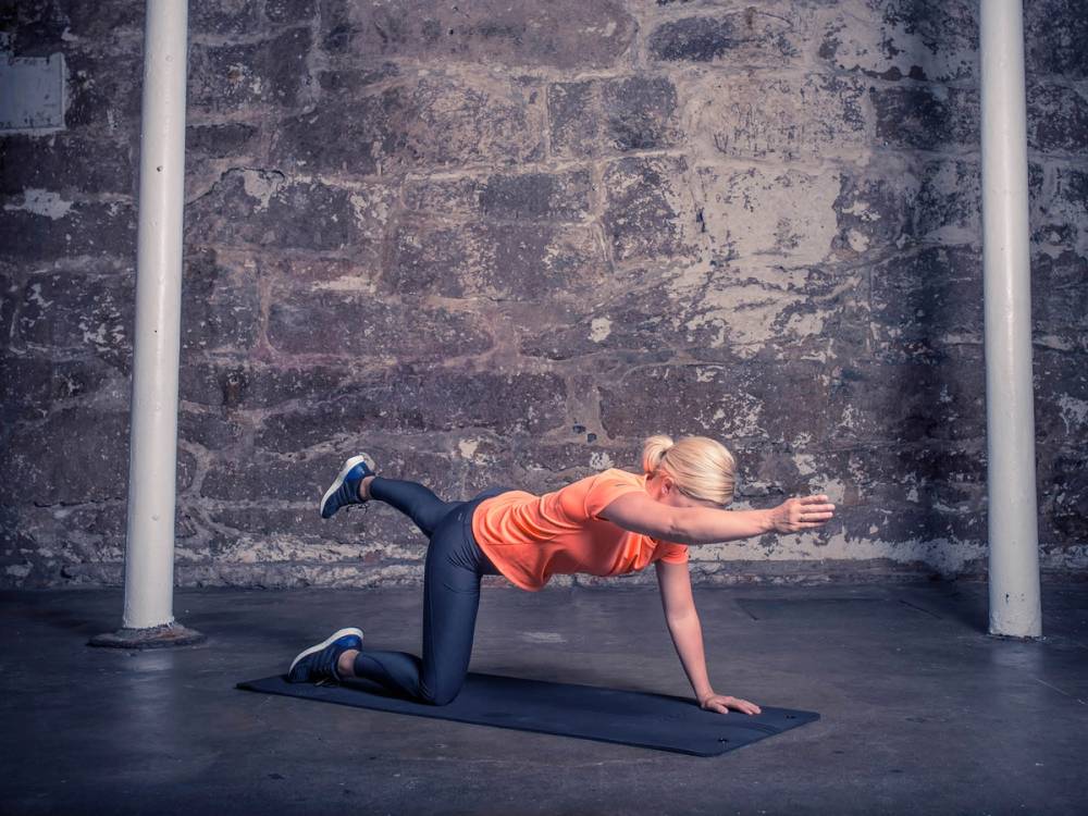 En kvinne i en rosa treningstøy gjør en øvelse på gulvet i et treningsrom med en murvegg i bakgrunnen.