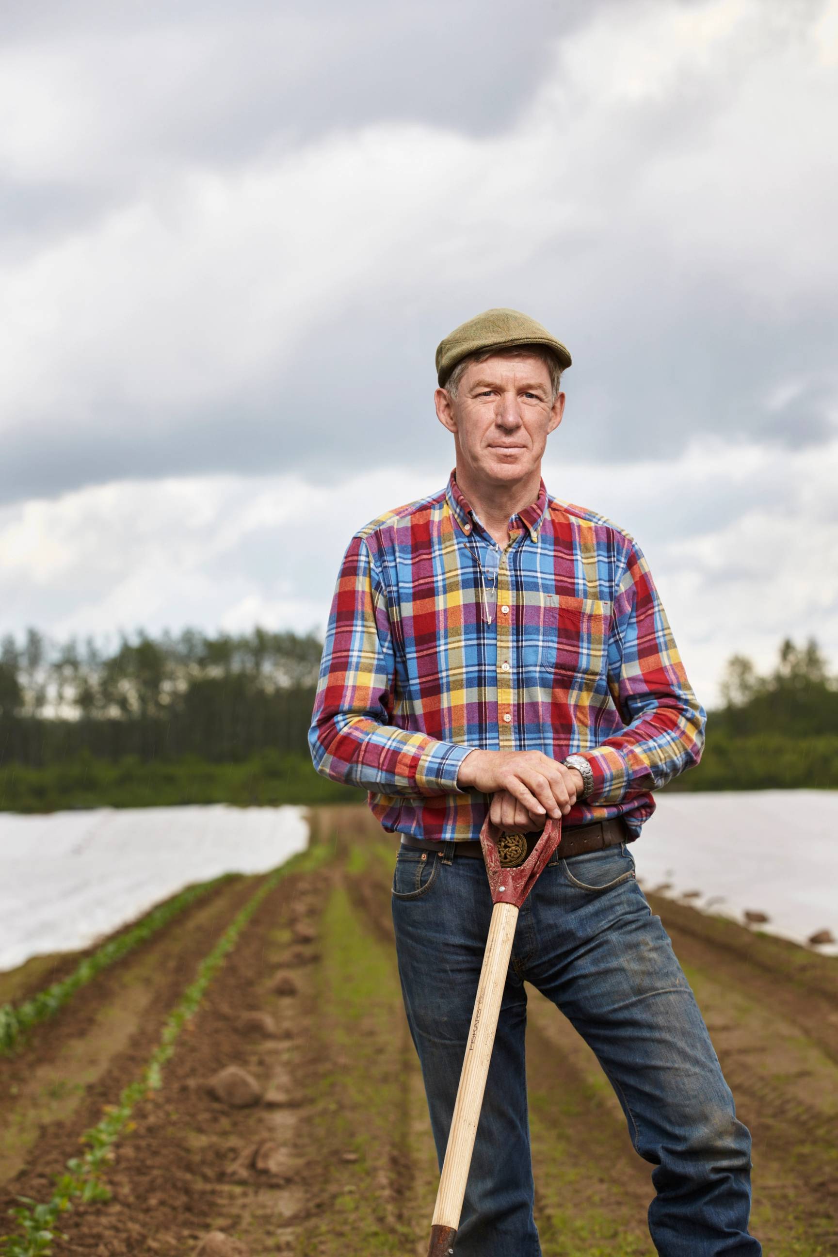 En person står i en jordbruksåker med en spade, iført en fargerik skjorte og en beige caps, med grønne planter og skyer i bakgrunnen.