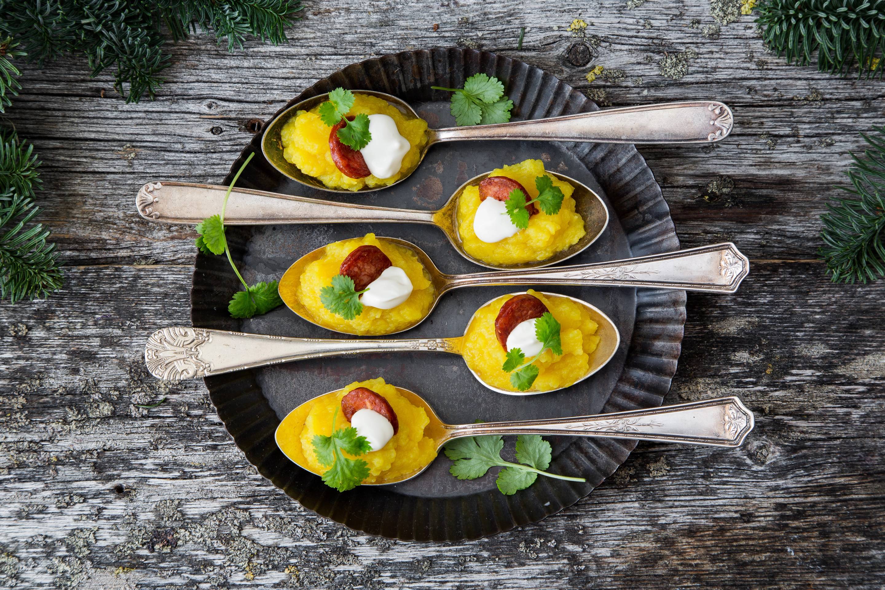 Fem små porsjoner med kålrabihorizo servert på skjeer på en svart tallerken med grønt garnityr, plassert på en treoverflate.