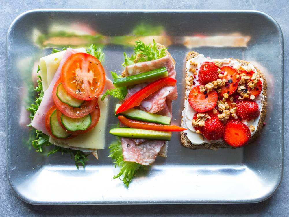 To brødskiver med pålegg, en med grønnsaker og skinke, og en med jordbær og granola, på en metallplate.