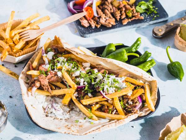 En gyrosrull med svinekjøtt, pommes frites, grønnsaker og saus på et fat, med tilbehør som jalapeños og pommes frites i bakgrunnen.