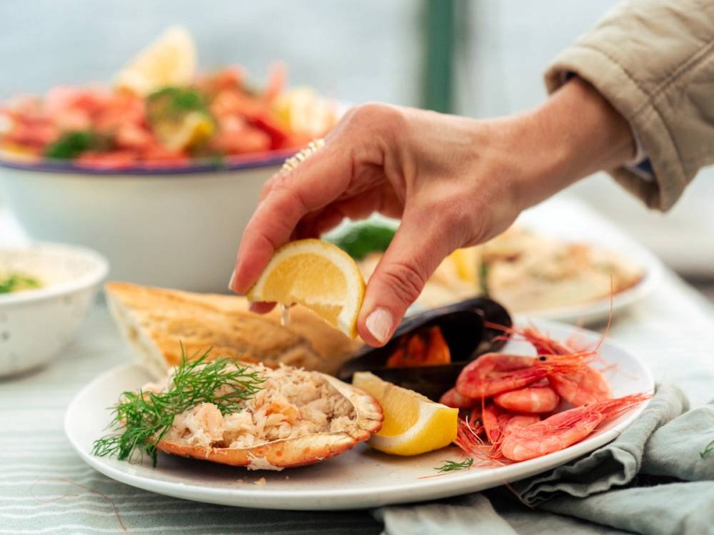 En person legger sitron på en tallerken med sjømat, inkludert reker og fisk, på et bord med flere retter.