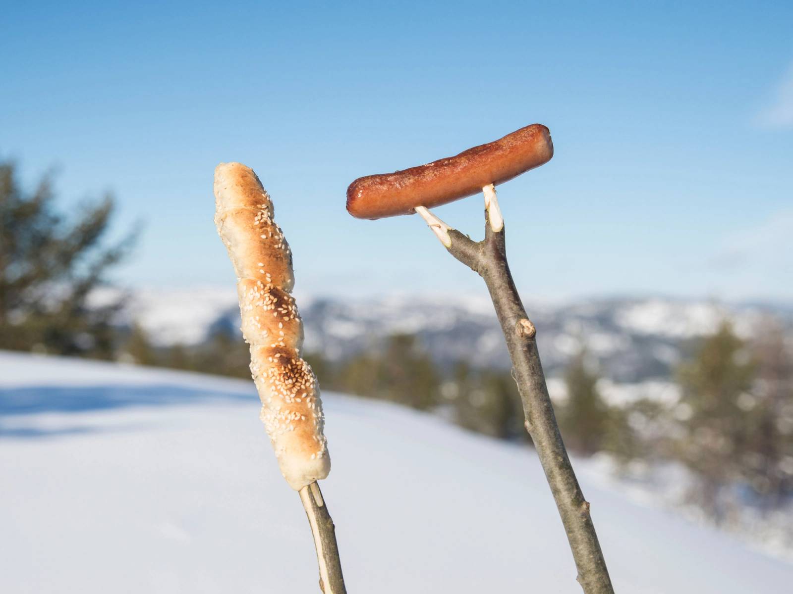 To pølser på spyd holdt opp i luften mot en snødekt fjellandskap, med klar blå himmel i bakgrunnen.