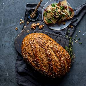 Et stort, ferskt brød med frø på toppen ligger på en svart klut, med en tallerken med smørbrød og grønnsaker i bakgrunnen.