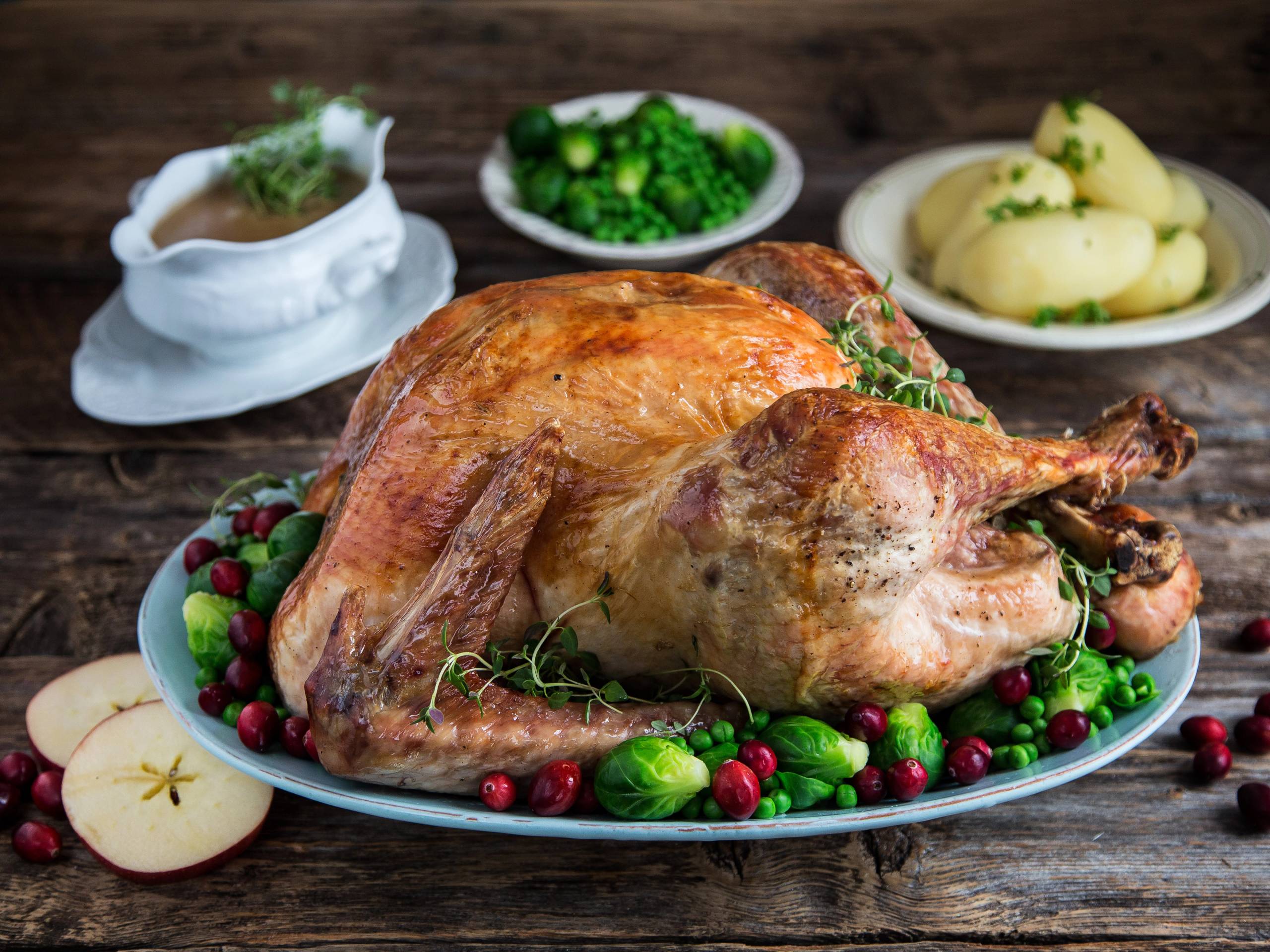En stekt kalkun på et serveringsfat omgitt av bær og grønnsaker, med poteter og saus i bakgrunnen.