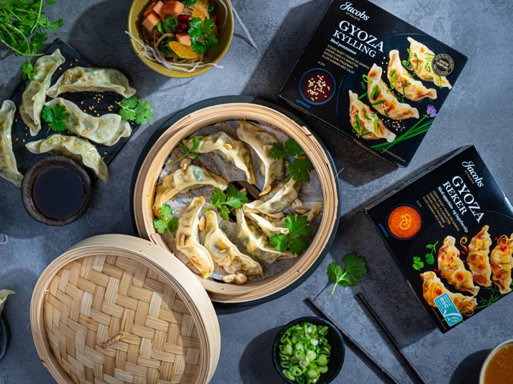 Diverse pakker med ferdigmat og en bambusdampkorg med dumplings, omgitt av små skåler med saus og grønnsaker, på et mørkt bord.