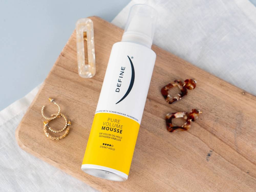 En flaske med hårmousse plassert på et trebord sammen med et par øreringer og en hårklips, med en hvit teksturert bakgrunn.