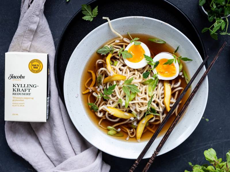 En bol med nudler, halverte egg og grønnsaker i en soyasaus, med spisepinner på siden, og en pakke med Jacobs Kylling Kraft Redusert ved siden av.