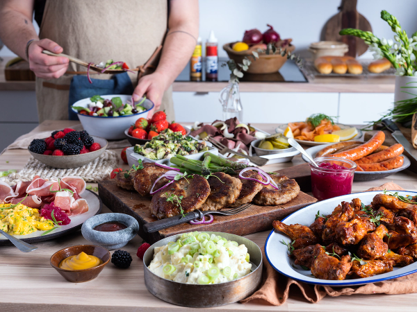 Et koldtbord med ulike typer kjøtt, salater, brød og frukt på et kjøkkenbord.