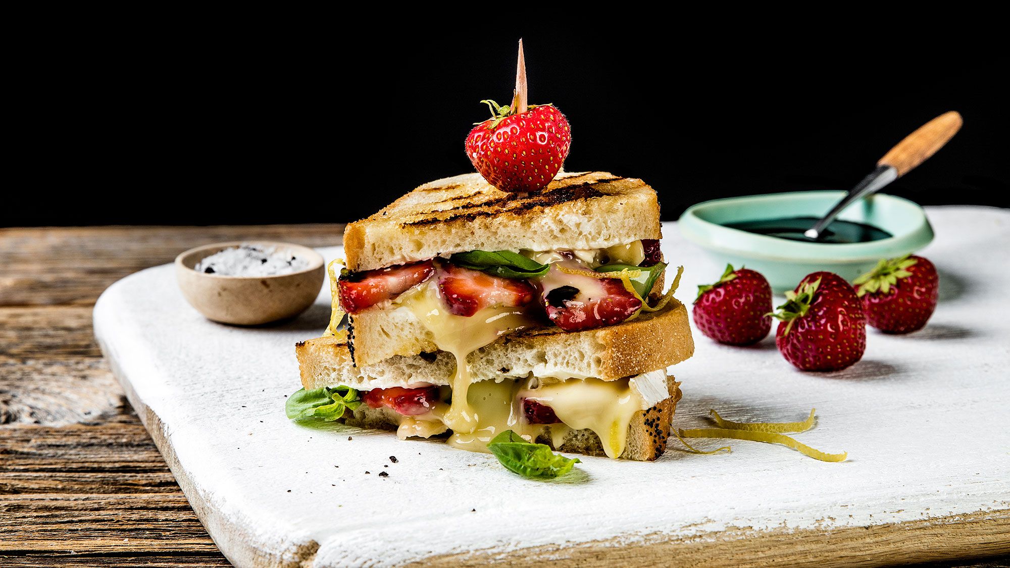 Ostesmørbrød med økologisk brie, jordbær og basilikum på en treplate med små skåler ved siden av.