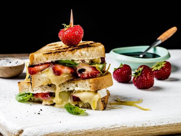 Ostesmørbrød med økologisk brie, jordbær og basilikum på en treplate med små skåler ved siden av.