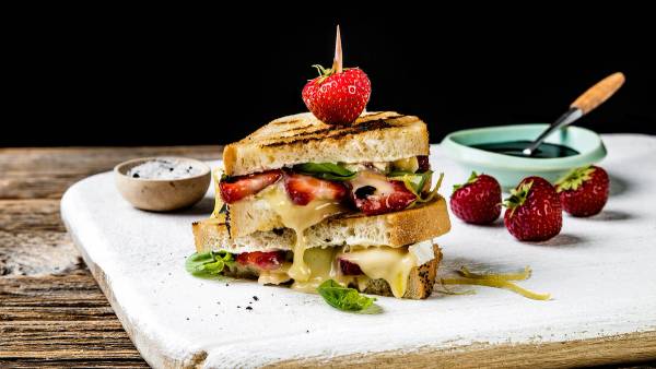 Ostesmørbrød med økologisk brie, jordbær og basilikum på en treplate med små skåler ved siden av.