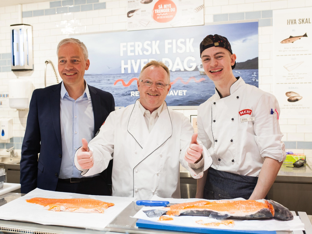 To menn i kokkeklær står bak en disk med fersk laks i en butikk eller fiskebutikk, og viser tommelen opp.