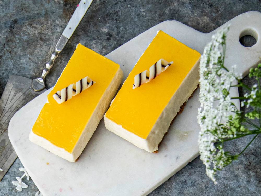 To ostekaker med gul glasur og dekorative sjokoladestenger på toppen, plassert på et hvitt skjærebrett med blomster i nærheten.