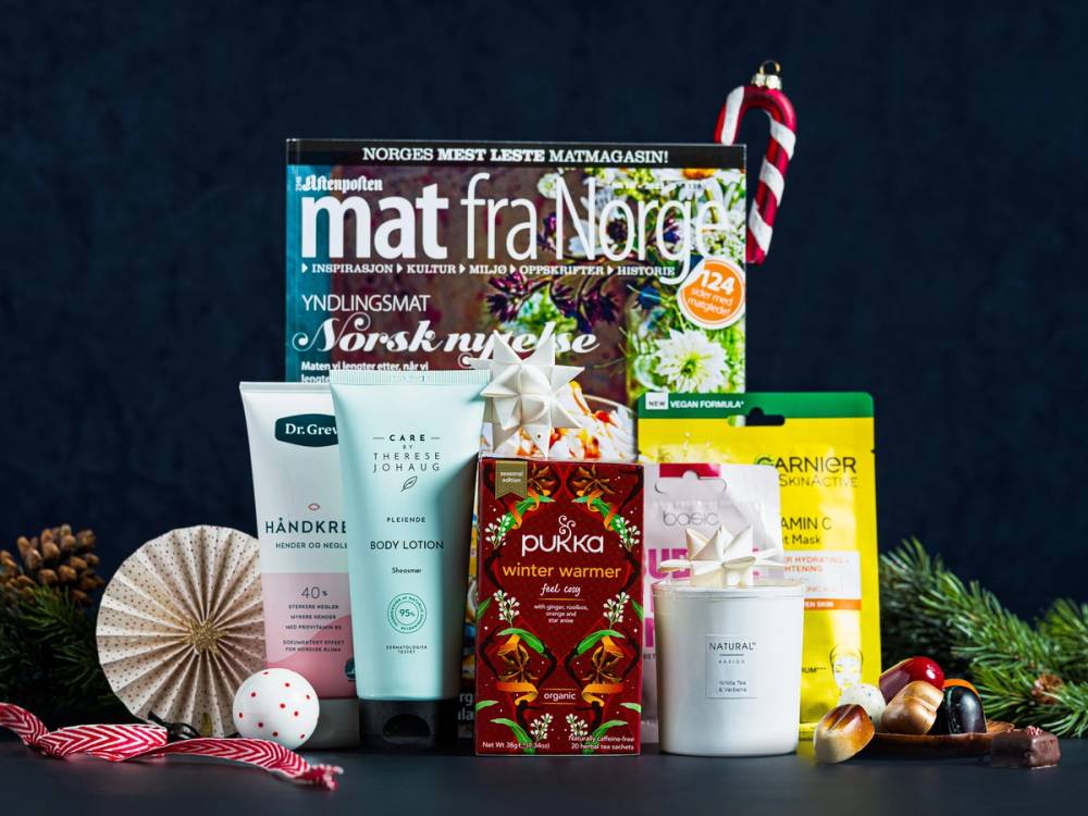 Ulike matgaver og julepynt på et svart bord, inkludert en julekrans, pepperkaker og julegaver, med en julehilsen i bakgrunnen.