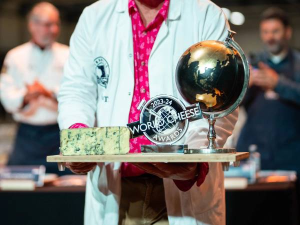 Person i hvit frakk holder en treplate med ost, en liten jordglob og en pris, med flere mennesker i bakgrunnen.