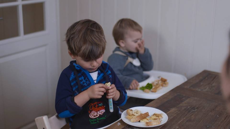 To barn sitter ved et bord og spiser, med en tallerken med mat foran dem, i et rom med hvite vegger og dører.