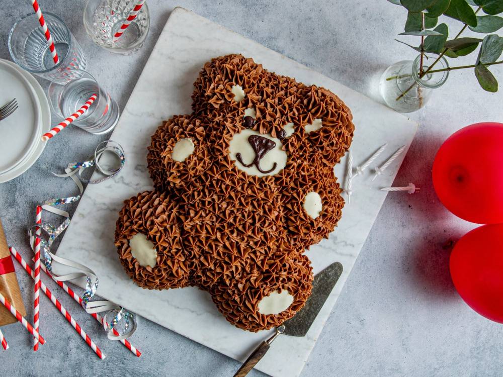 En bamsekake dekorert med sjokoladestrøssel og hvite detaljer, plassert på et hvitt underlag med julepynt rundt.