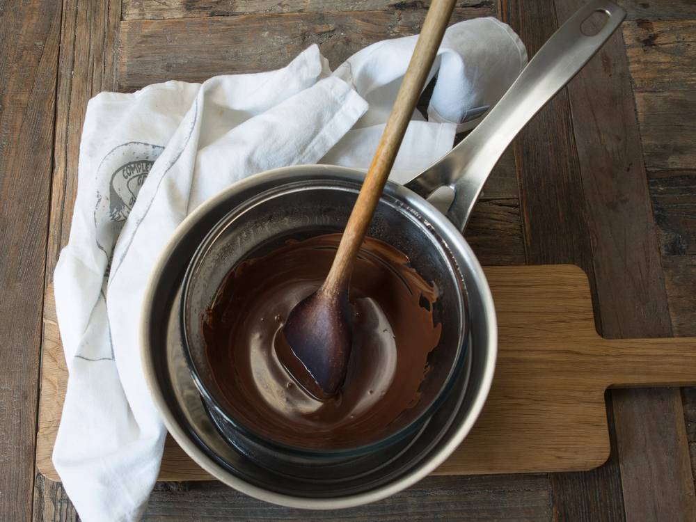 Smeltet sjokolade i en kjele med en tresleiv, plassert på et trebord med en hvit klut ved siden av.