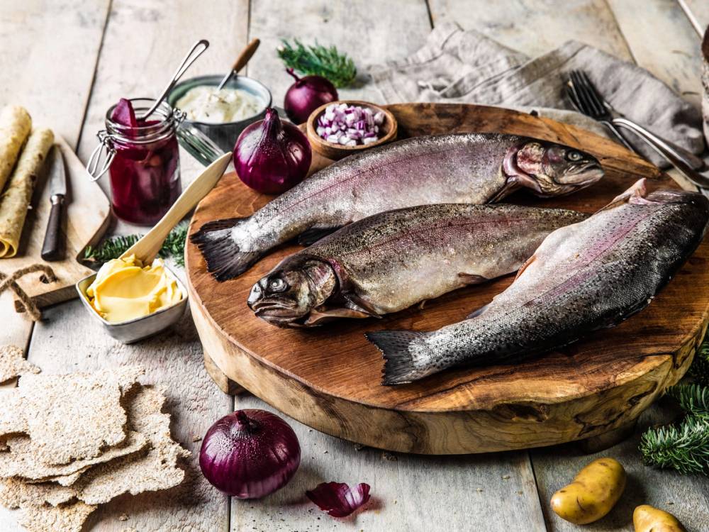 To helle rakfisk ligger på et rundt trefat omgitt av små glass med tilbehør og rødløk, på et rustikt trebord.