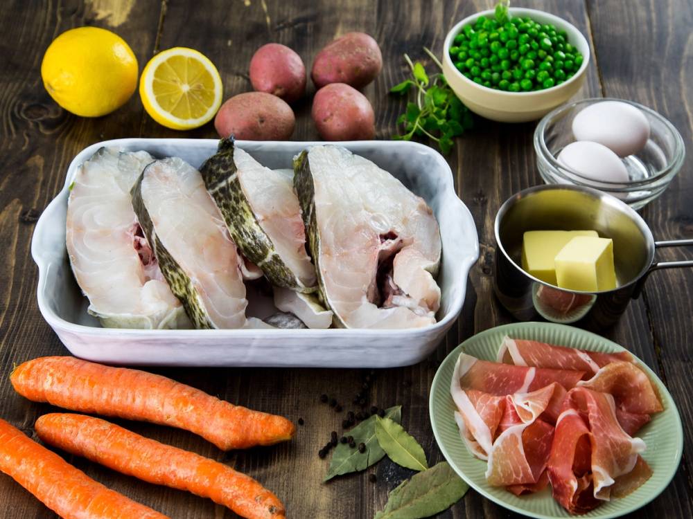 Skiver av skrei med tilbehør som poteter, gulrøtter, sitron, egg, smør, og spekeskinke på et bord.