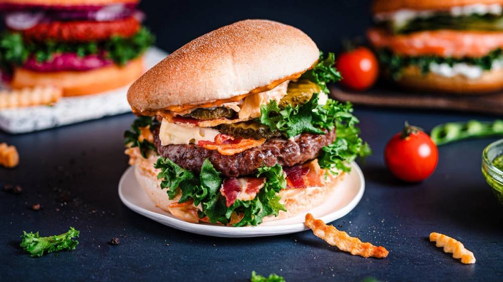 En saftig burger med kjøttdeig, salat og tomat på en hvit tallerken, med flere andre burgere i bakgrunnen.