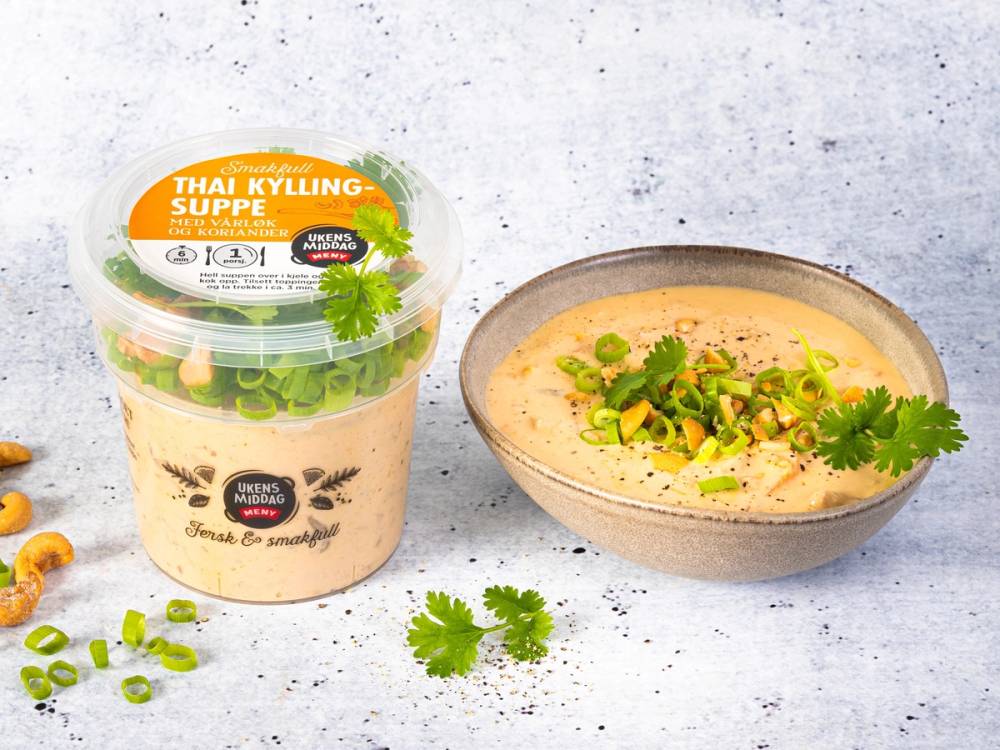 En kopp thai kyllingsuppe med grønnsaker og koriander, og en plastbeholder med suppe ved siden av på en lys bakgrunn.