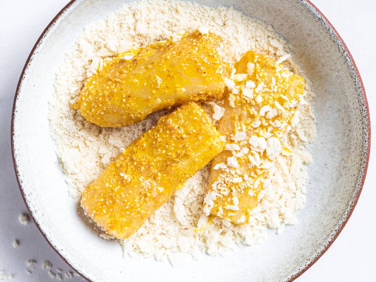 To stykker ferdigpanert kylling legges i en skål med mel og revet ost, klare til steking.