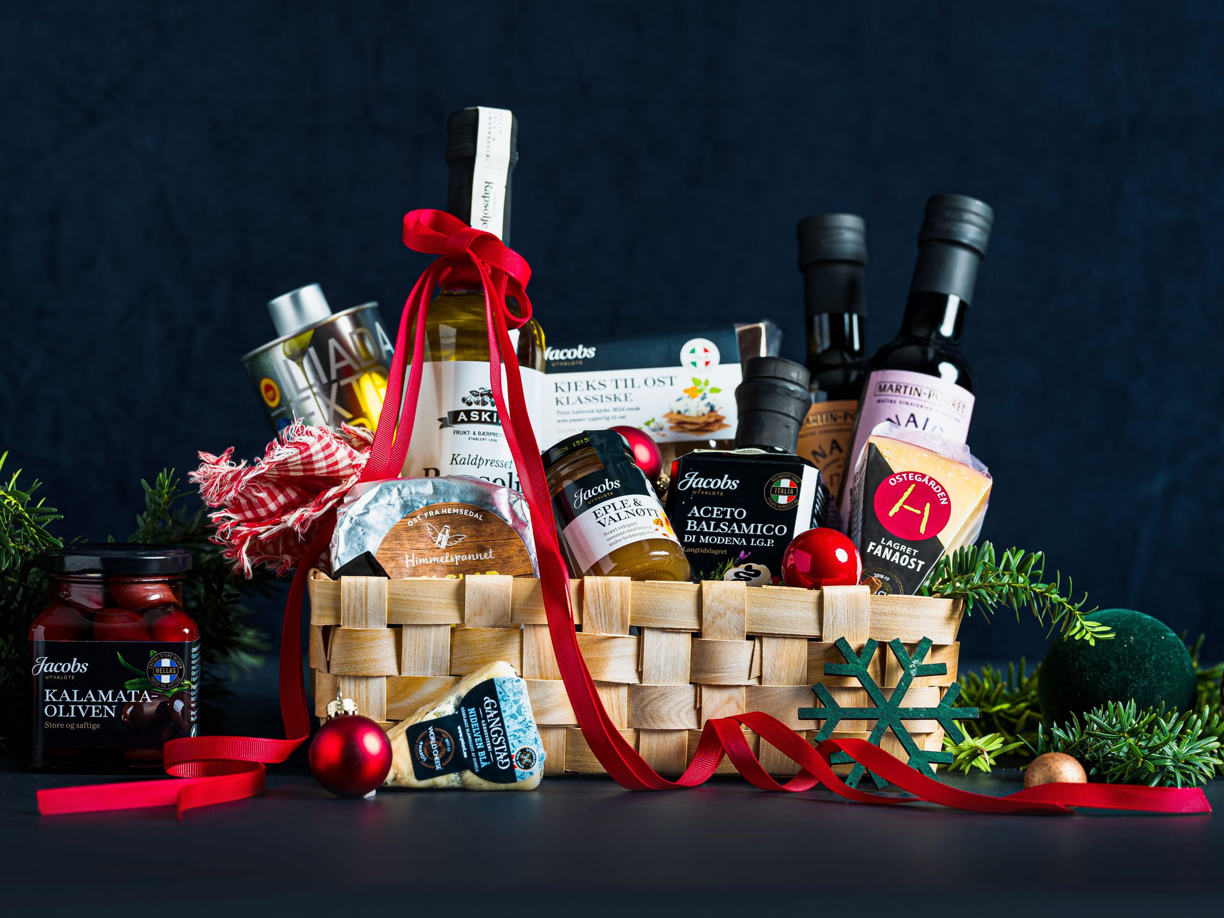 En julegavekurv fylt med matvarer som oljer, syltetøy, og små juledekorasjoner, omgitt av røde ornamenter og grønt til julestemning.