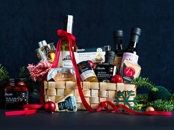 En julegavekurv fylt med matvarer som oljer, syltetøy, og små juledekorasjoner, omgitt av røde ornamenter og grønt til julestemning.