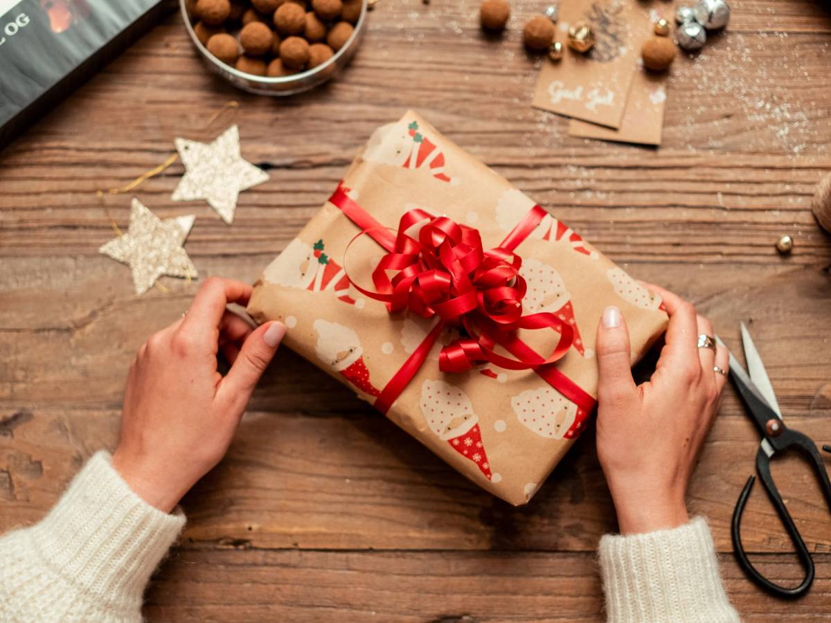 Person som pakker inn en gave med julepapir og rød sløyfe på et treverkbord, omgitt av julepynt og gaver.