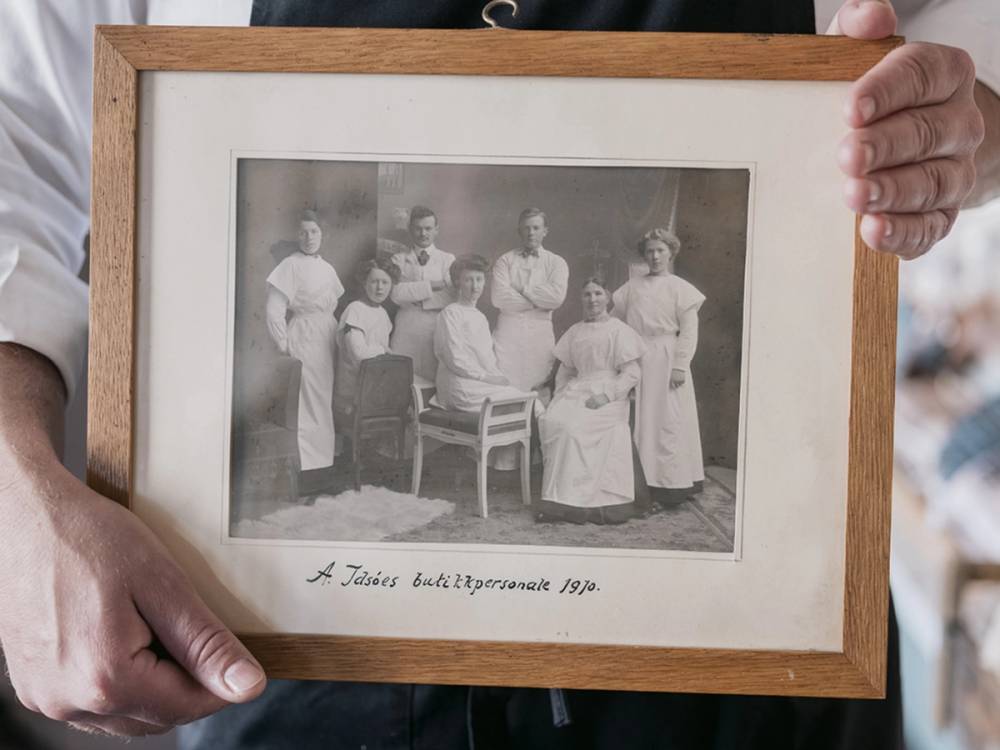 En person holder opp et svart-hvitt familiebilde i en ramme, med flere voksne og barn som poserer sammen, trolig fra 1920-tallet.