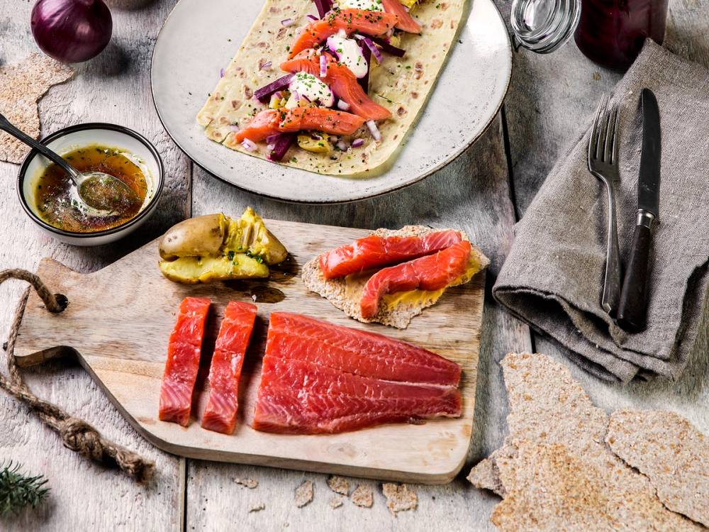 Rakfisk servert med flatbrød, rømme, og tilbehør på et bord med bestikk og glass.