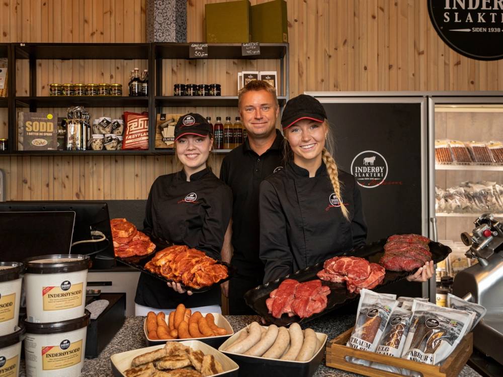 Tre ansatte i slakteri eller kjøttbutikk står bak disken med ulike typer kjøtt og pølser, med tre personer i uniform og en bakgrunn av trepanel og produkter på hyller.