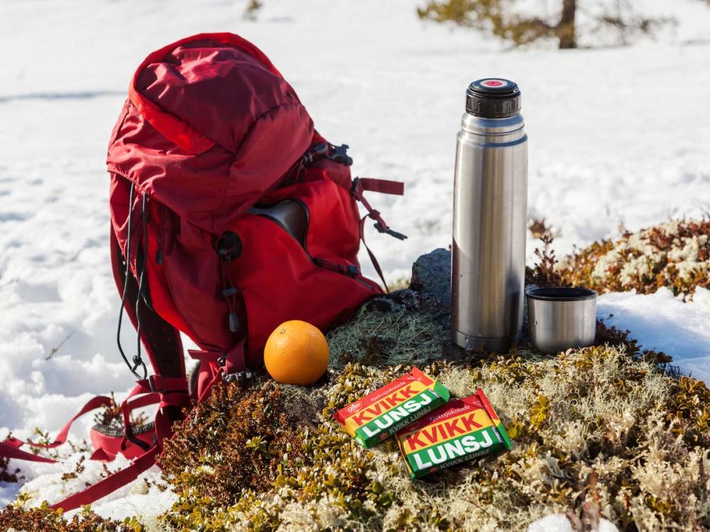 En rød tursekk med en termos, en liten kopp, en appelsin og en pakke Kvikk Lunsj ligger på snødekket ute i naturen.