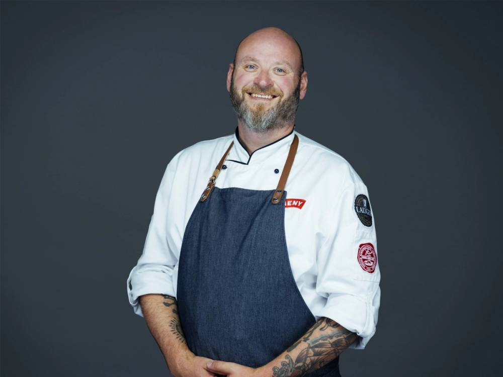 En person iført kokkeuniform og forkle, med tatoveringer på armene, står mot en mørk bakgrunn.