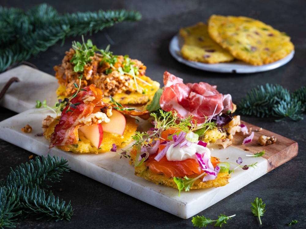 Saftige potetkaker med ulike pålegg som skinke, rømme og urter på en skjærebrett, med flere potetkaker i bakgrunnen.
