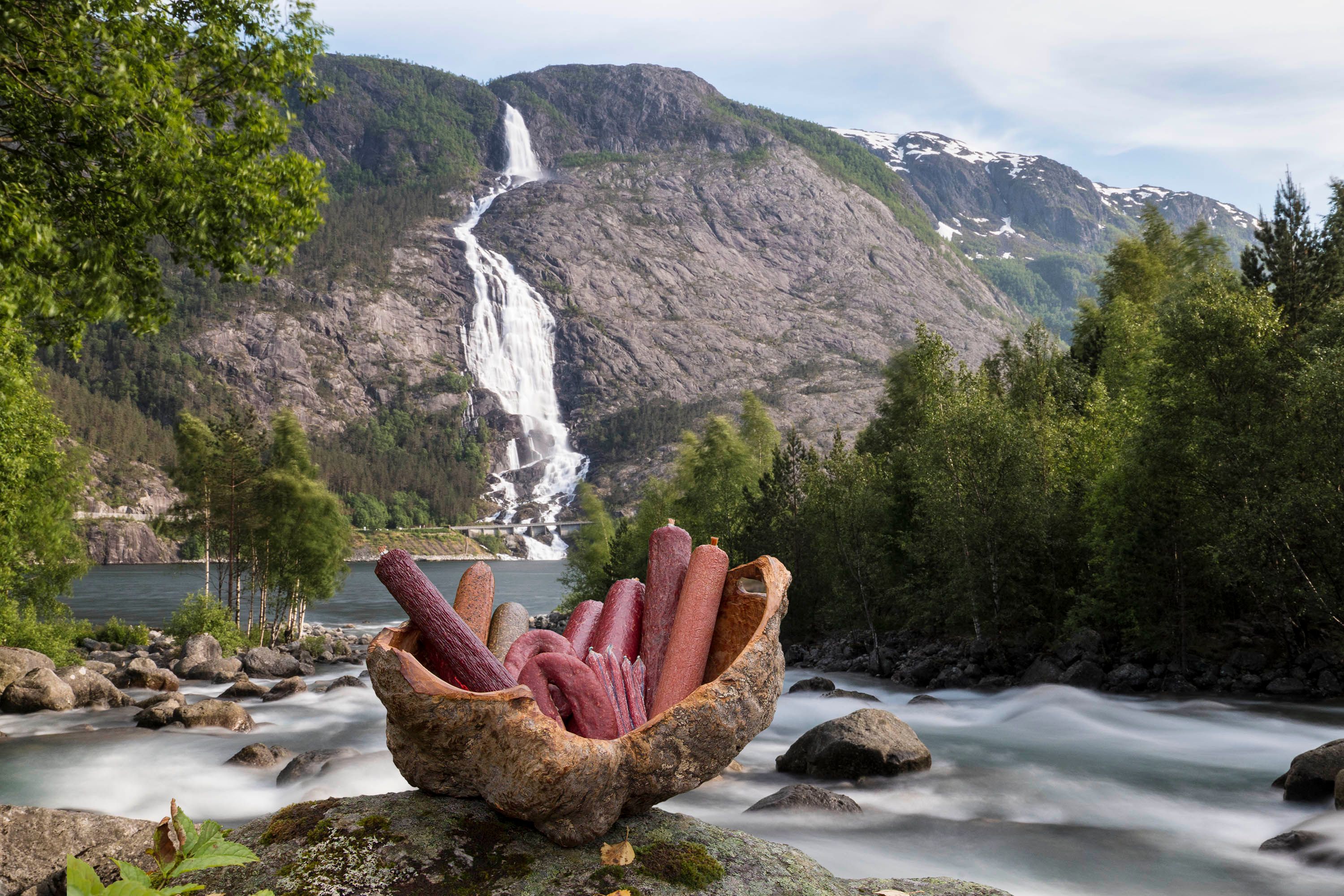 En fjellandskap med en foss som faller nedover en bratt fjellside, omgitt av grønne trær og en elv i forgrunnen med en kurv fylt med pølser.