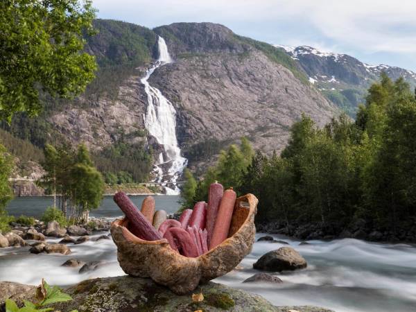 En fjellandskap med en foss som faller nedover en bratt fjellside, omgitt av grønne trær og en elv i forgrunnen med en kurv fylt med pølser.