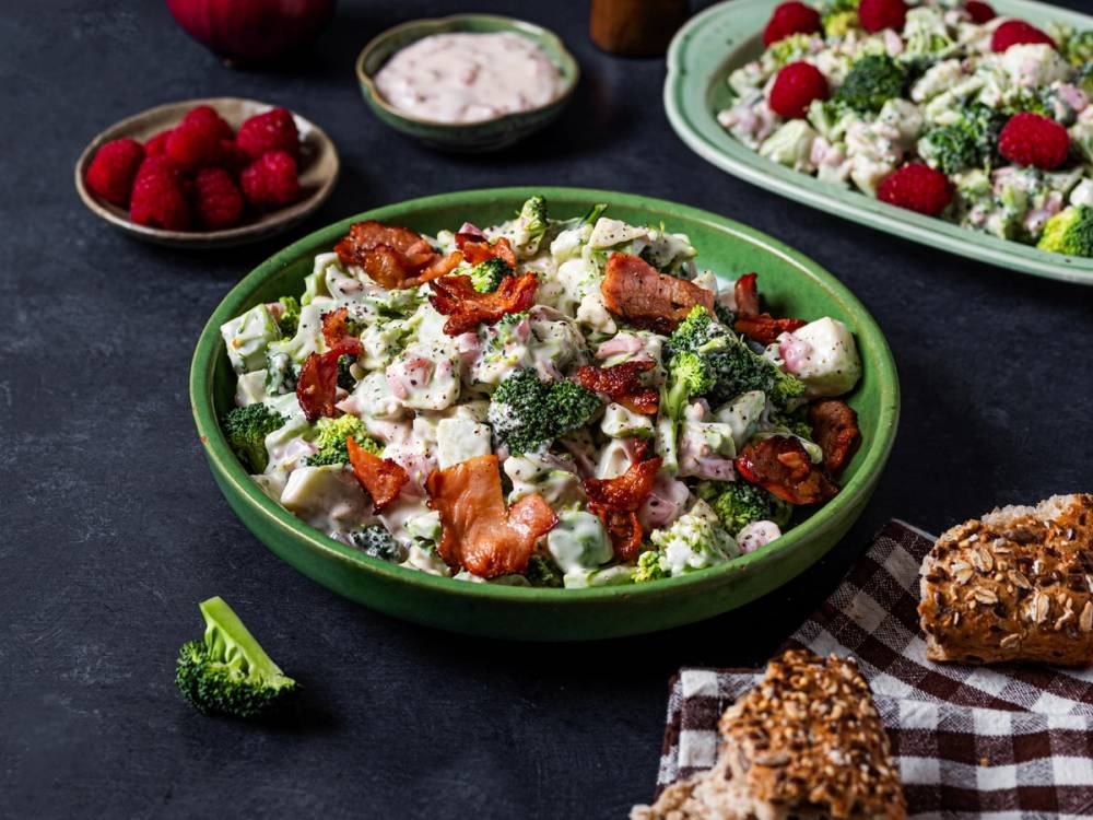 En salat med brokkoli, tomater, ost og bacon i en grønn bolle, med flere salater og snacks rundt på bordet.