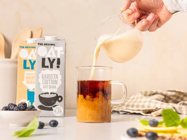 En person heller melk i en kopp med kaffe, med melkekartong og blåbær på bordet, på et kjøkkenbord.