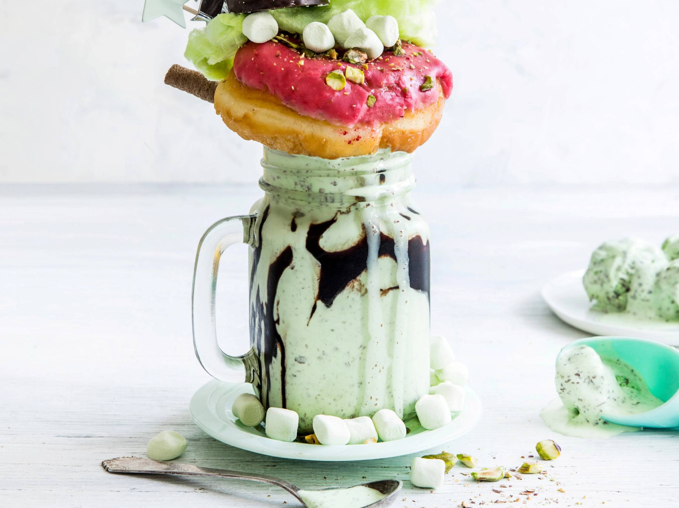 En milkshake med iskrem i en glass med sjokoladesaus og pyntet med små godteribiter og en kjeks, med flere iskremkuler og en skje ved siden av.