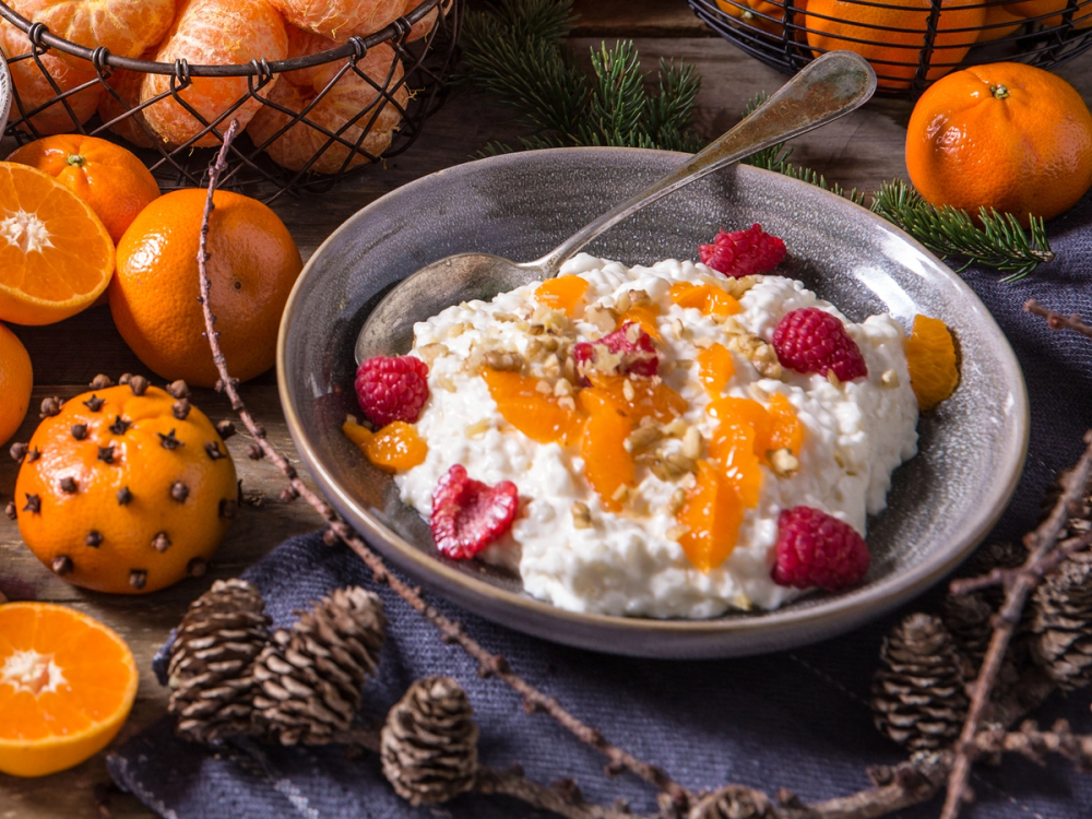 Riskrem med klementinbåter og bringebær i en grå skål, omgitt av julenisser, klementiner og kongler på et julebord.