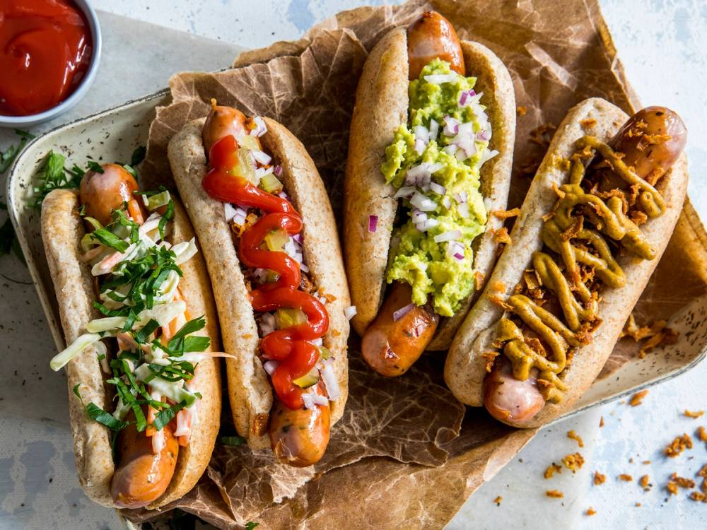 Tre pølser i brød med ulike toppings som grønnsaker og saus, på papir med en liten skål med ketchup ved siden av.