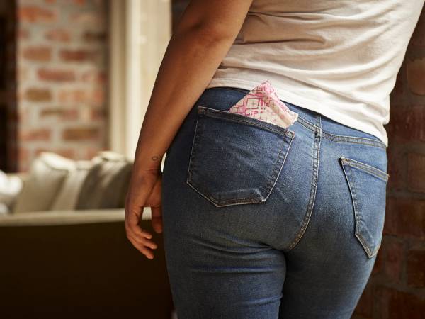 En person står med ryggen mot kamera, iført blå jeans og en hvit t-skjorte, med en rosa og hvit mønstrete lomme i baklommen.
