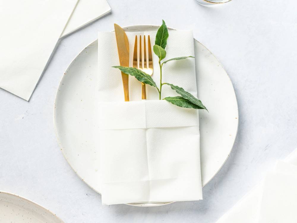 En hvit serviett brettet med en liten grønn plante og en gullfarget gaffel oppå, plassert på en hvit tallerken.
