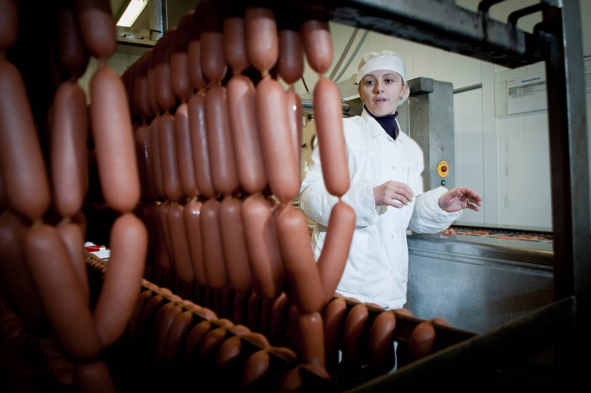 En person i hvit kokkekjole og blå forkle står bak en vegg laget av pølser i en matbutikk eller på et marked.