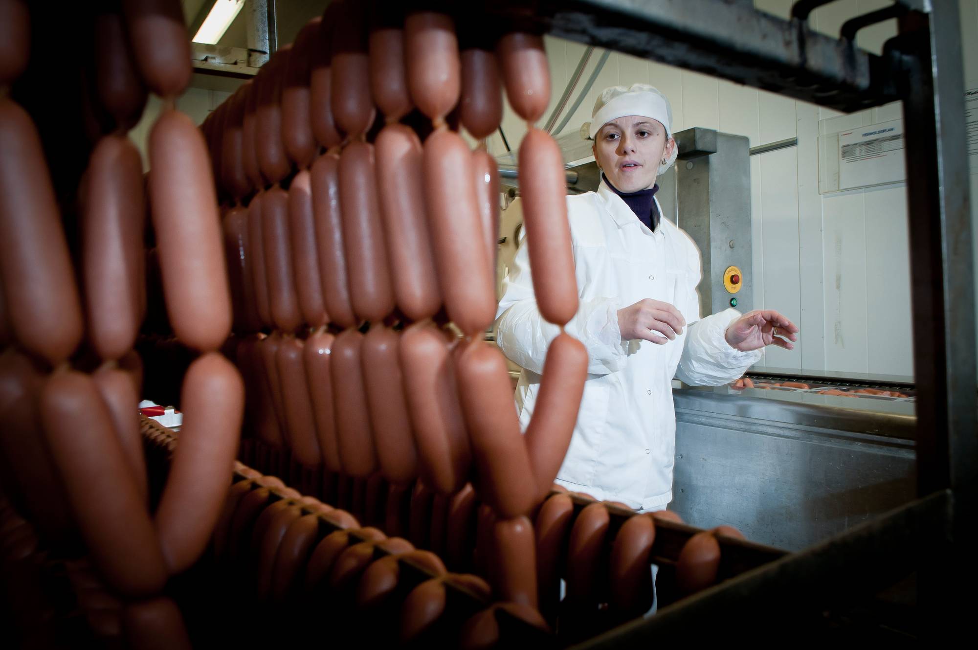 En person i hvit kokkekjole og blå forkle står bak en vegg laget av pølser i en matbutikk eller på et marked.