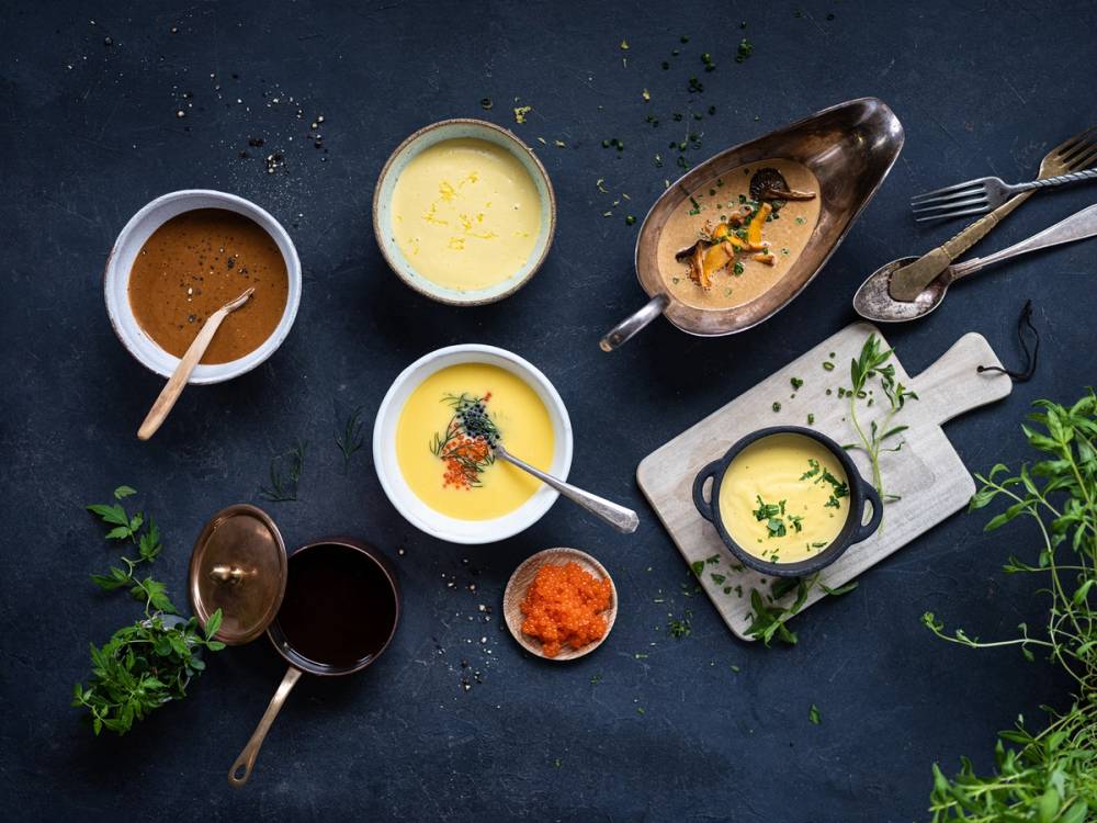 Ulike sauser og kraft i små skåler og kopper på et mørkt bord, med urter og krydder rundt